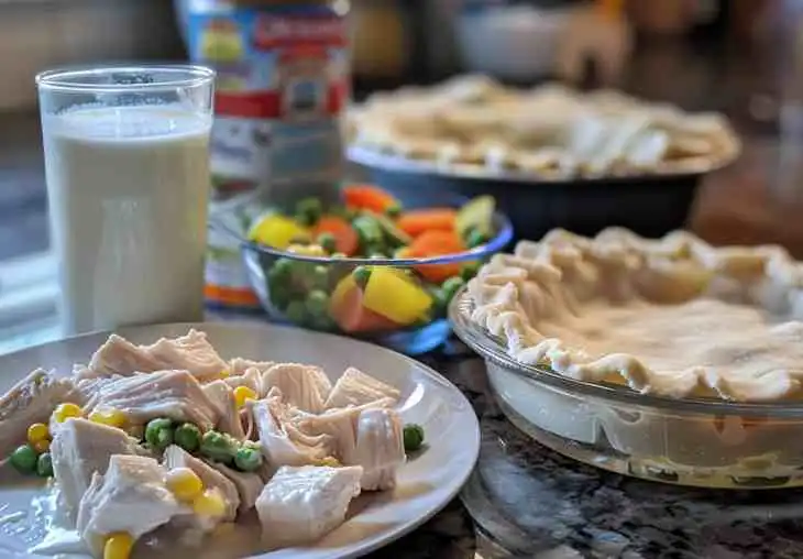 Ingredients for chicken pot pie including cream of chicken soup, milk, vegetables, and pie crust prepared on a kitchen counter.