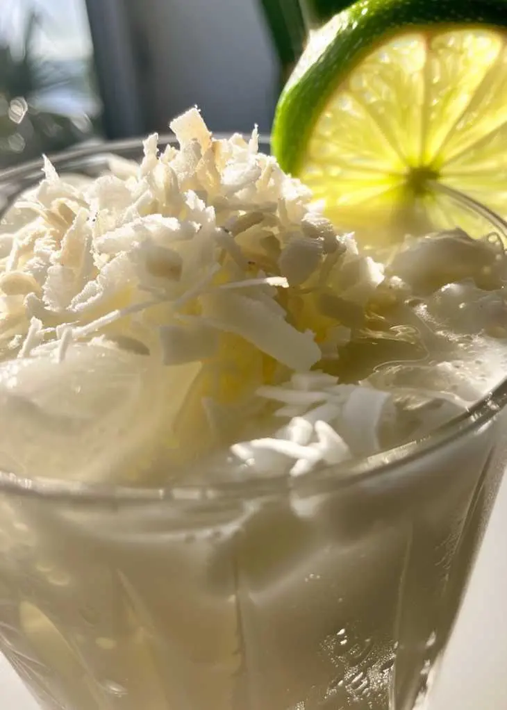 Close-up of creamy lime and coconut drink with shredded coconut topping and a lime slice in natural sunlight.