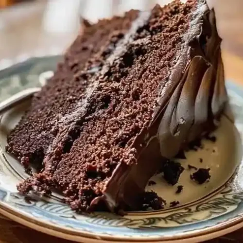 Moist triple chocolate cake slice on a plate with creamy chocolate frosting and natural daylight, perfect for homemade dessert inspiration.
